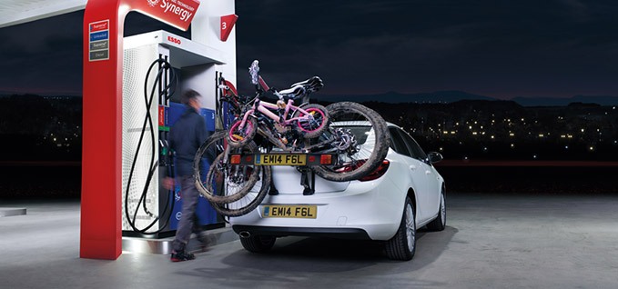 A white car with 3 bikes on the back is in an Esso station at night