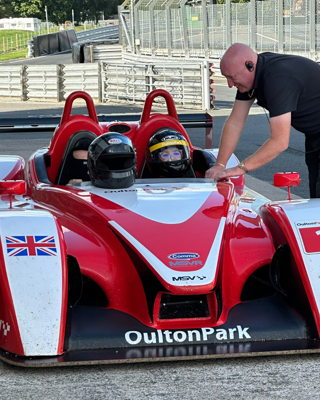 Two helmeted participants seated in a red sports prototype car at Oulton Park as an instructor speaks to them.