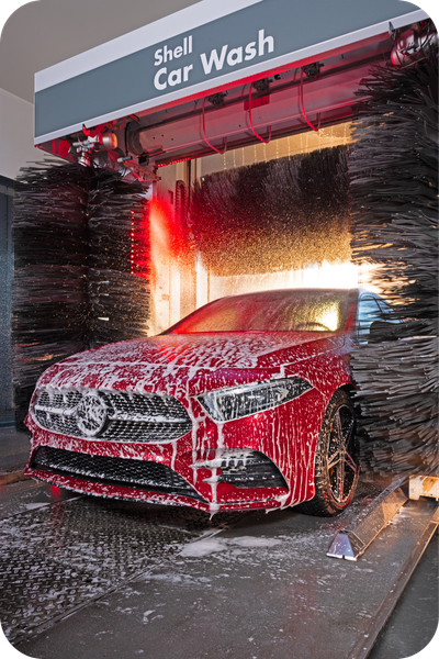 Red company car covered in foam while being cleaned inside an automatic Shell Car Wash.